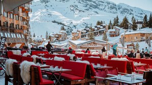 A snow-covered outdoor dining area with wooden tables and red chairs, surrounded by snow-covered buildings and mountains in the background. - Résidence Pierre & Vacances Premium L'Amara (Avoriaz)
