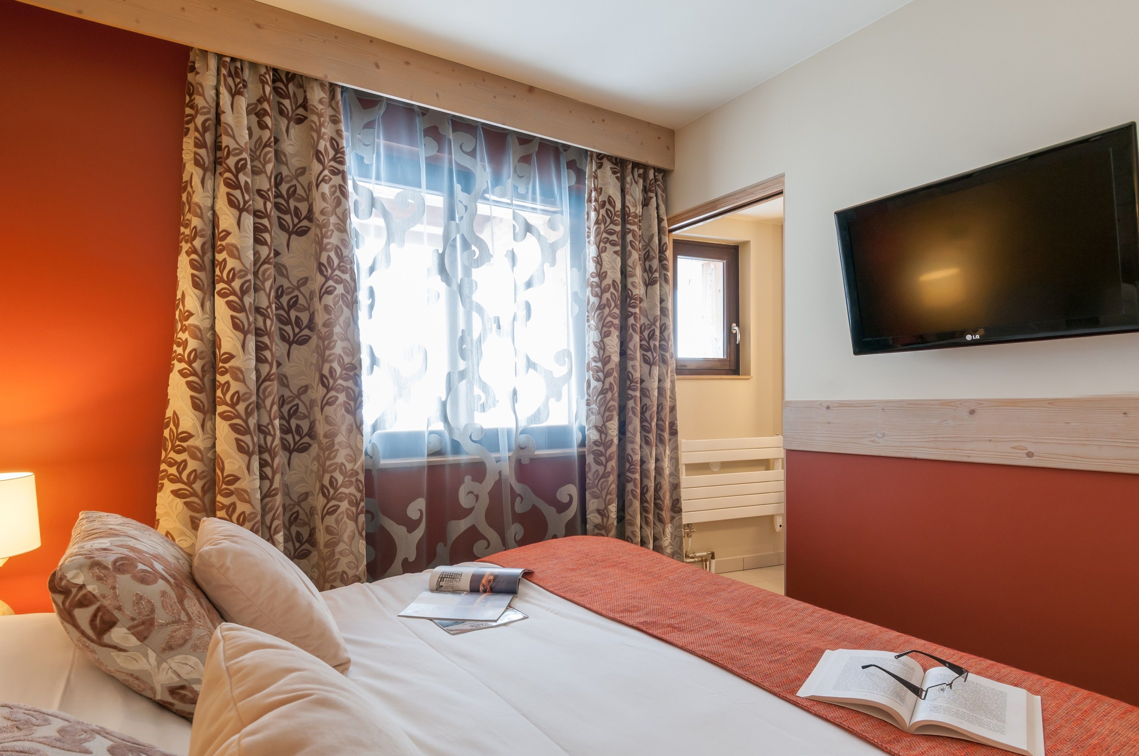 A hotel room with a bed, a flat-screen TV mounted on the wall, and a window with curtains.