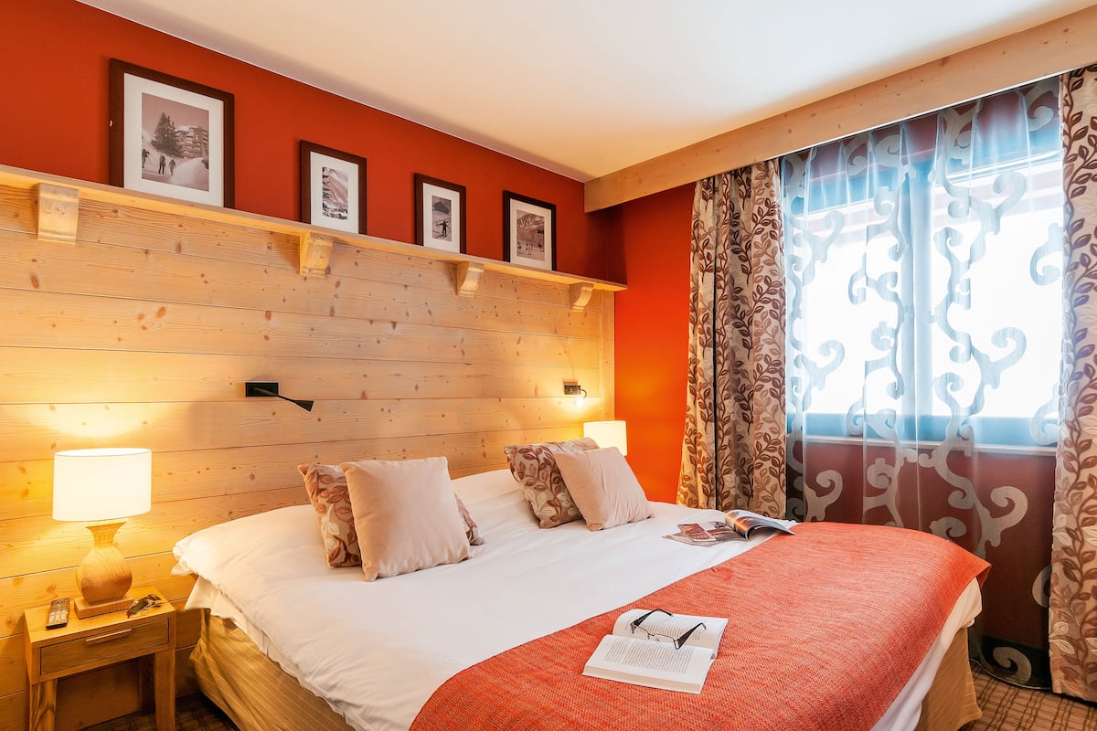 A hotel room with a wooden headboard, a bed with white bedding, a red throw blanket, and a nightstand with a lamp.