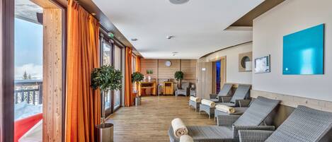 Vestíbulo de hotel con suelo de madera, ventanas amplias con cortinas naranjas, mostrador de recepción y sillones con toallas.