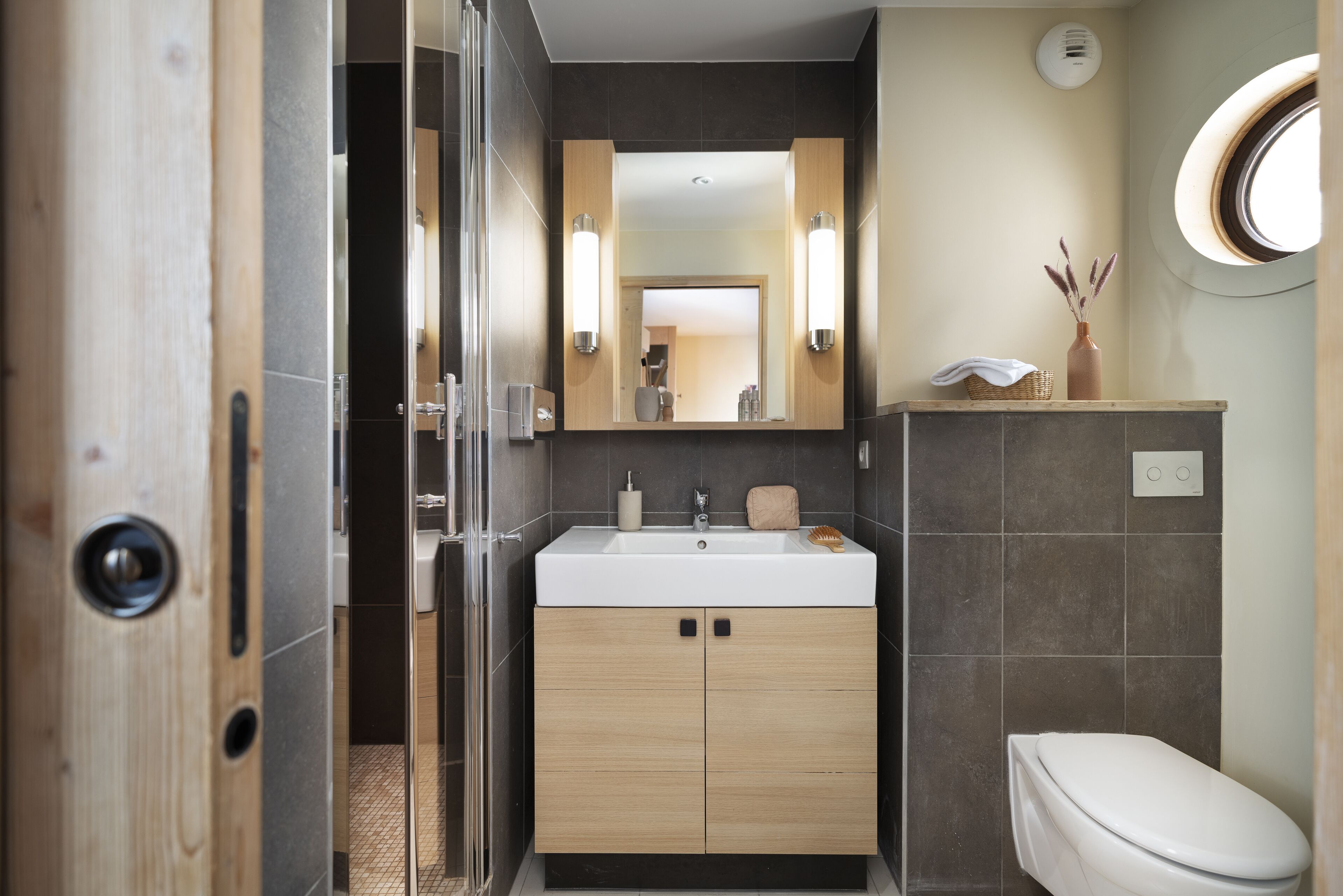 Un baño moderno con tocador de madera, un lavabo blanco y un ventanal redondo.