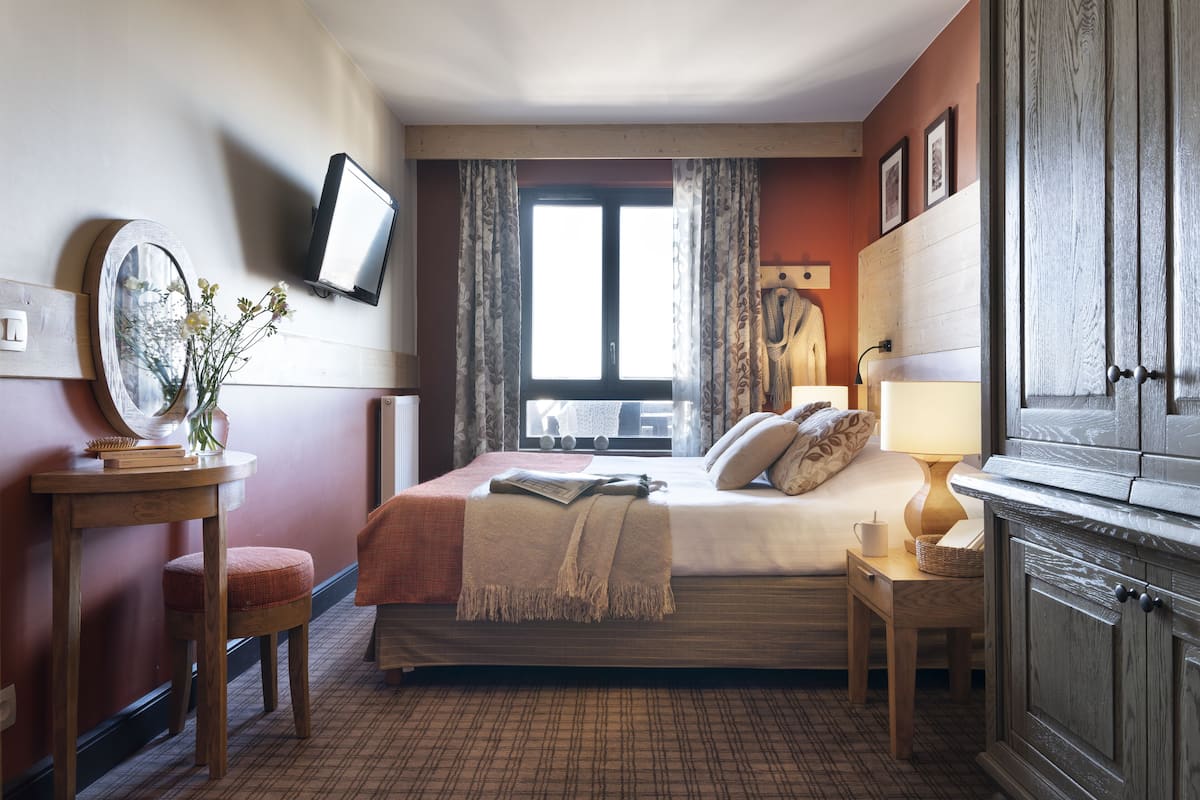 A hotel room with a bed, bedside table, lamp, TV, and a window with curtains.