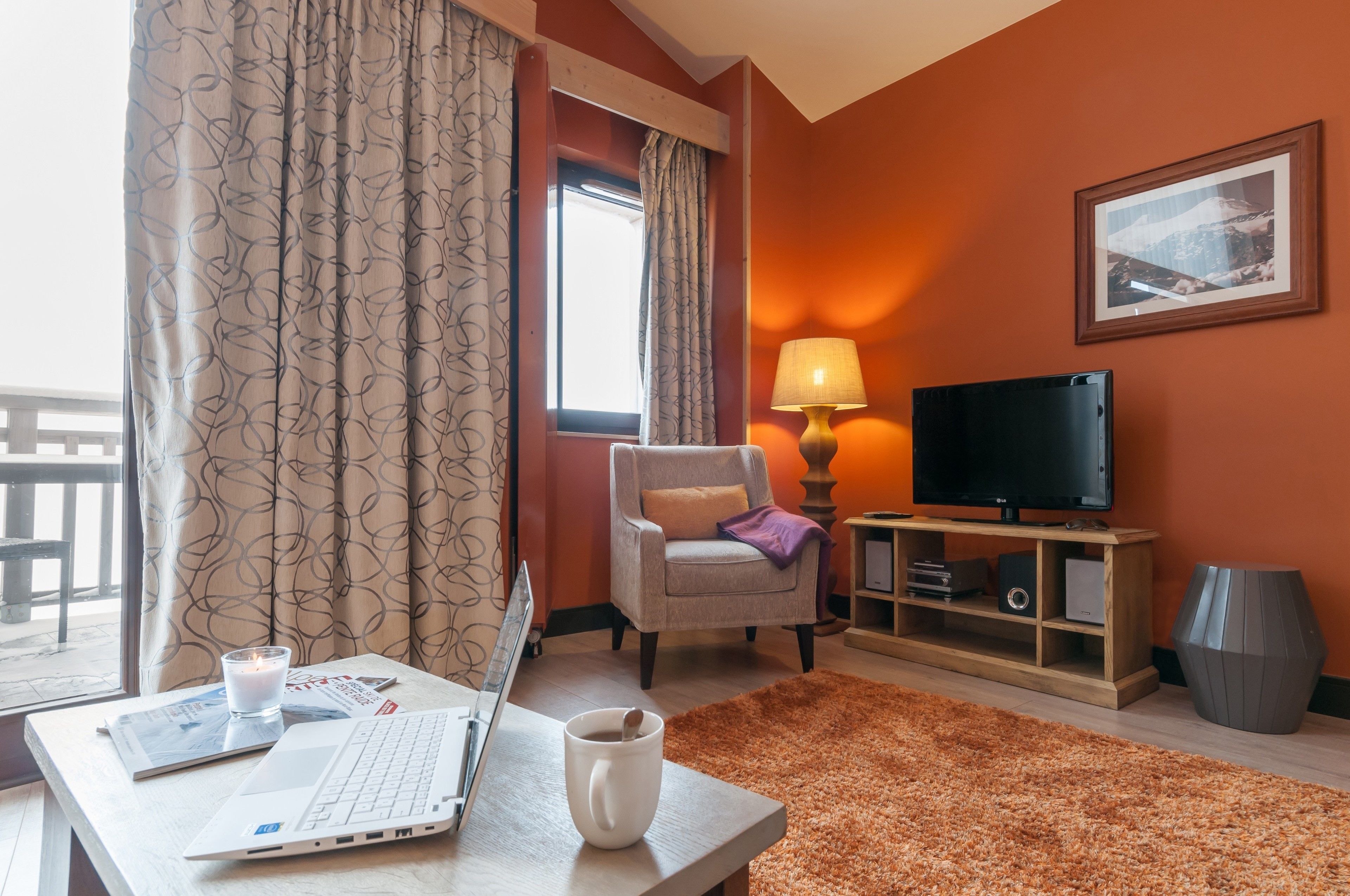 Habitación de hotel con una computadora portátil, una taza de café, un televisor, una silla, una lámpara y un cuadro en la pared.