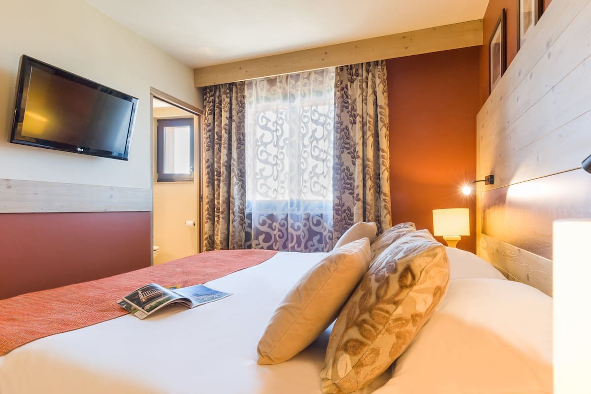 A hotel room with a bed, a flat-screen TV mounted on the wall, a window with patterned curtains, and a bedside lamp.