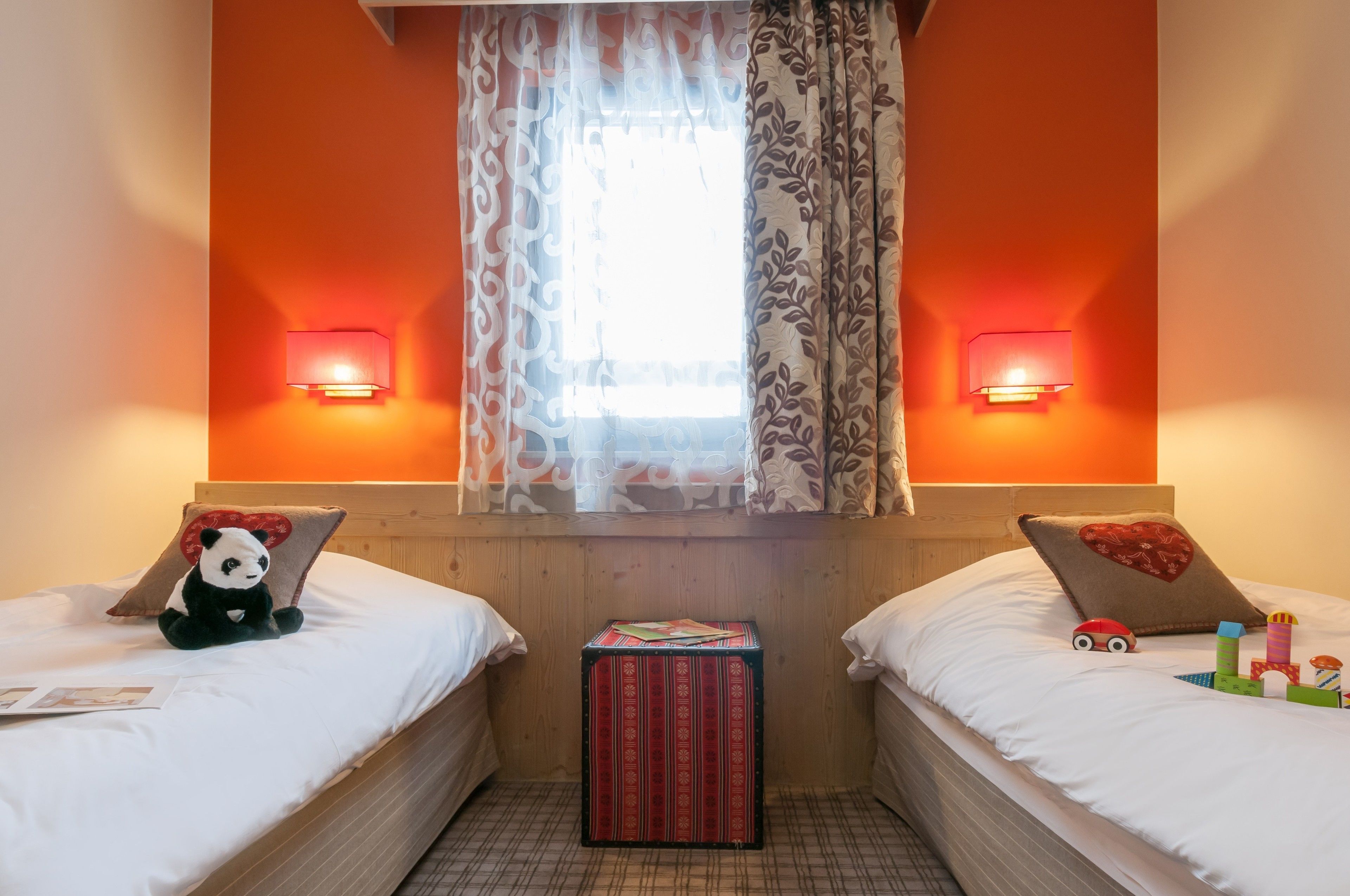 A hotel room with two beds, a window with patterned curtains, wall-mounted lamps, and a small table with a red box.