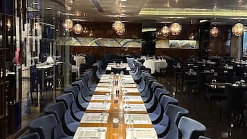 A long dining table with blue chairs, set for a meal, in a modern restaurant with a glass partition on one side.