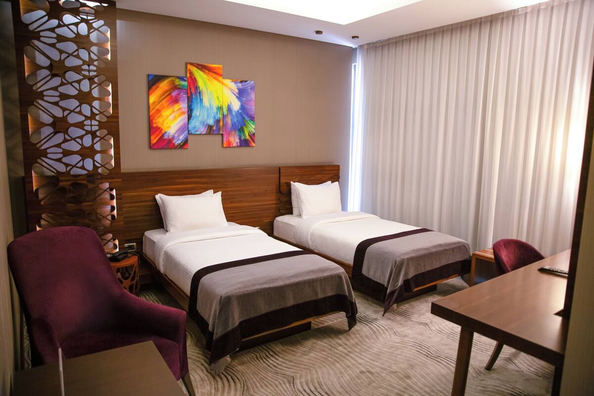 Habitación de hotel con dos camas, cabecero de madera, panel decorativo en la pared y un amplio ventanal con cortinas.