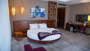 Une chambre d’hôtel avec un lit rond, un bureau, une chaise et un mur décoratif orné d’œuvres d’art abstraites.