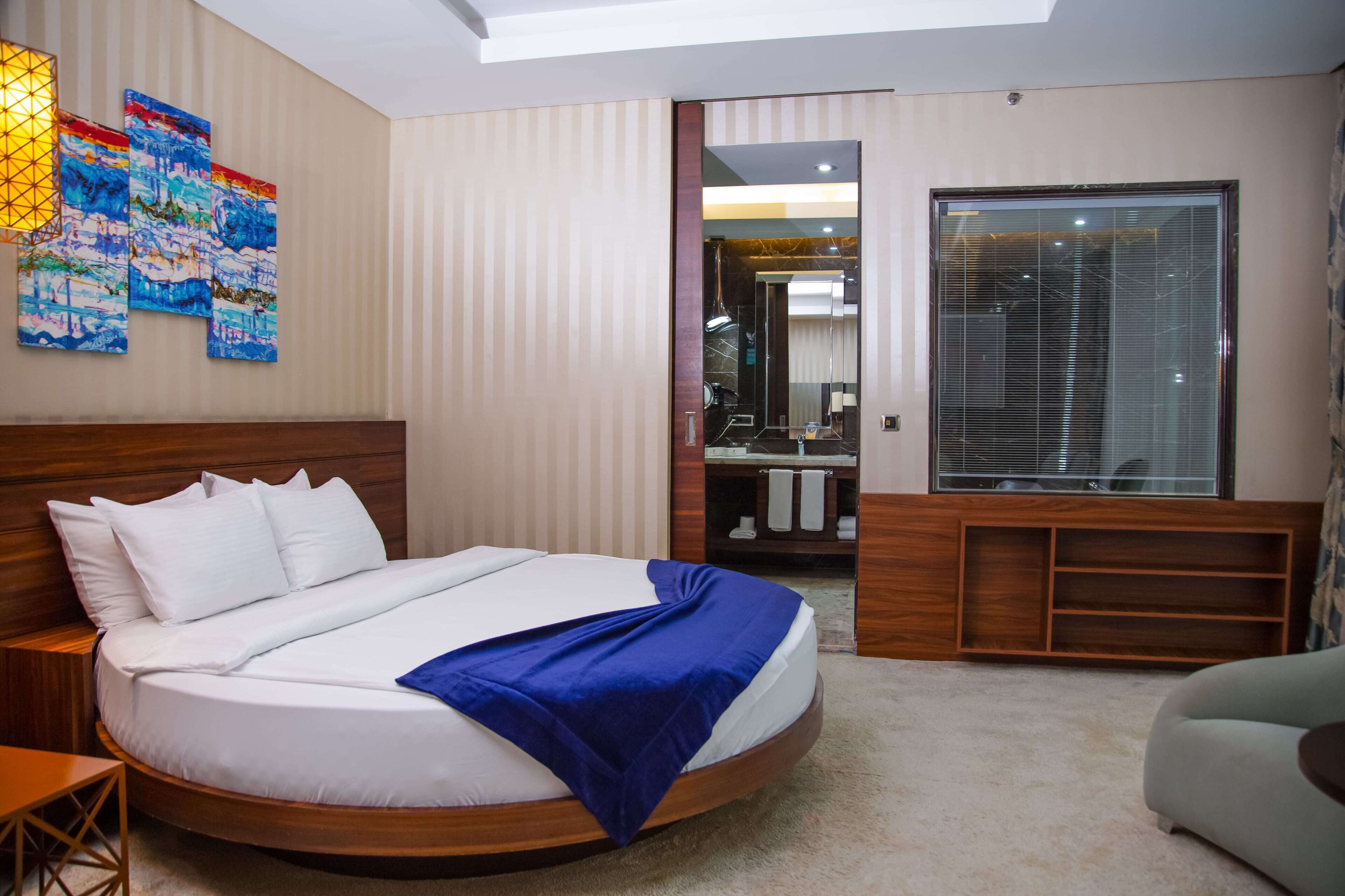 A hotel room with a large bed, a wooden headboard, a blue throw blanket, a wooden nightstand, a small table, a chair, a wall-mounted artwork, a mirror, a vanity with a sink, and a large window with blinds.