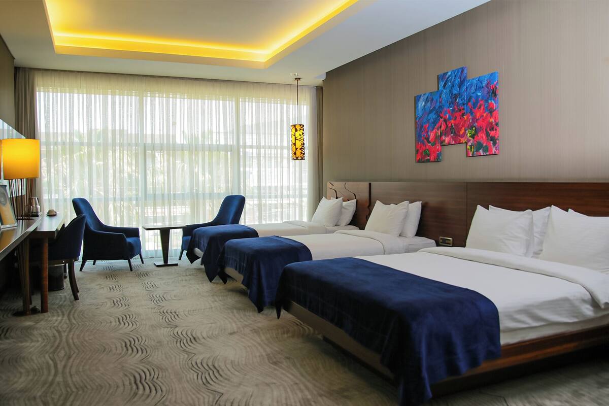 Habitación de hotel con dos camas, un escritorio, una silla y un ventanal con cortinas translúcidas.
