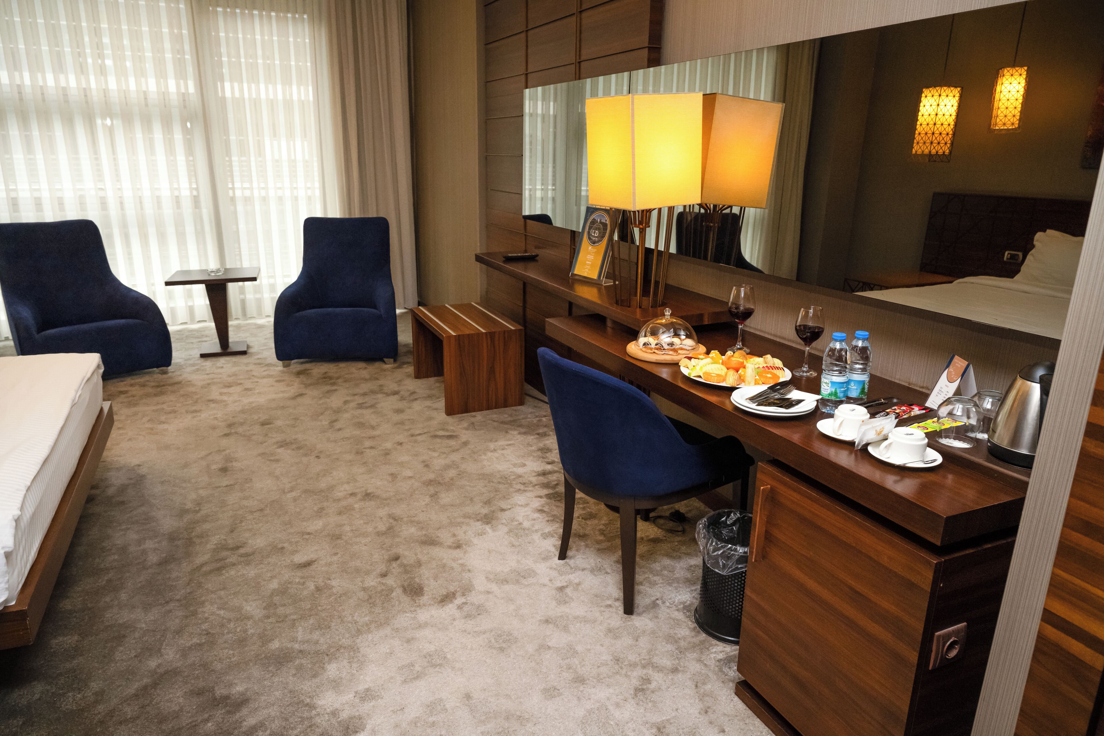 Una habitación de hotel con una cama, dos sillones azules, un escritorio de madera con una lámpara, una mesita y un minibar.