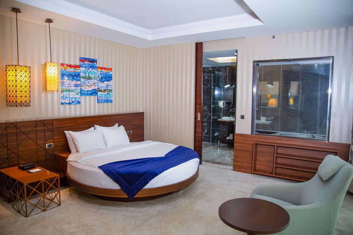 Habitación de hotel con una cama grande, una mesita de noche, una silla, un escritorio y un ventanal con persianas.