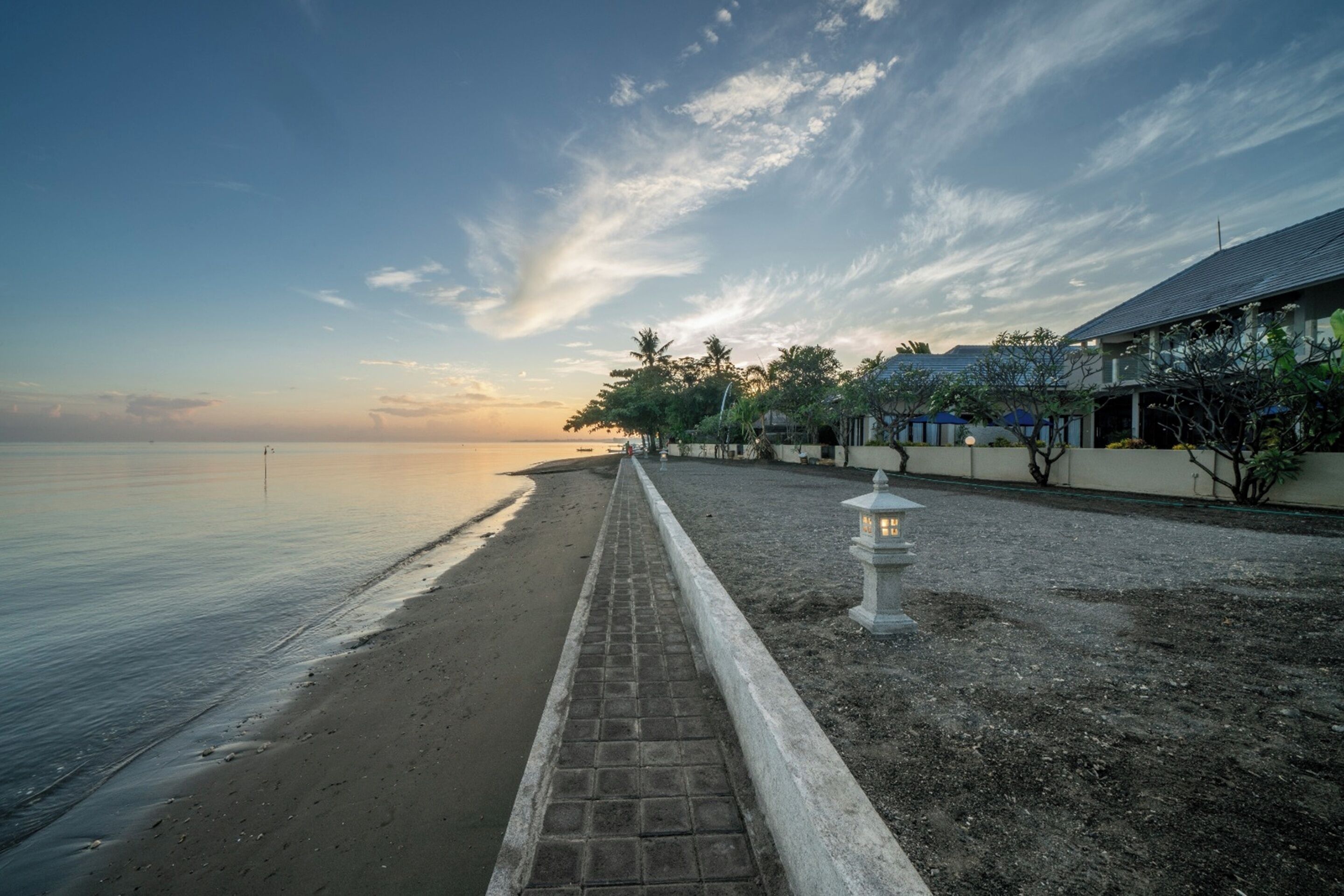 A beachside walkway with a brick path, a lamp post, and a building with a sloped roof in the background.