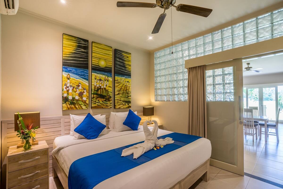 Ein Hotelzimmer mit einem großen Bett, einem Deckenventilator, Bildern an der Wand und Blick ins Freie.