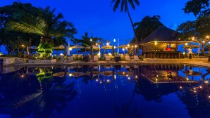 A nighttime resort pool area with lounge chairs and umbrellas, surrounded by palm trees and a building with a thatched roof. - The Lovina (Buleleng)