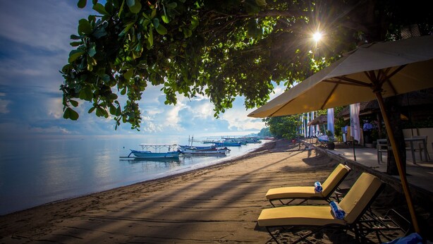 A beach with lounge chairs and umbrellas, boats in the water, and a clear sky. - The Lovina (Buleleng)
