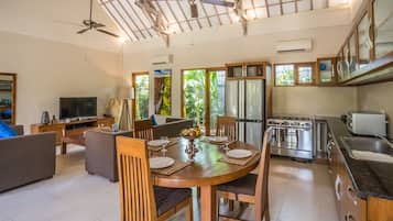 Ampla sala de estar com uma mesa de jantar de madeira preparada para quatro pessoas, rodeada por assentos confortáveis. Uma cozinha com eletrodomésticos modernos e uma grande janela com vista para o verde é visível.