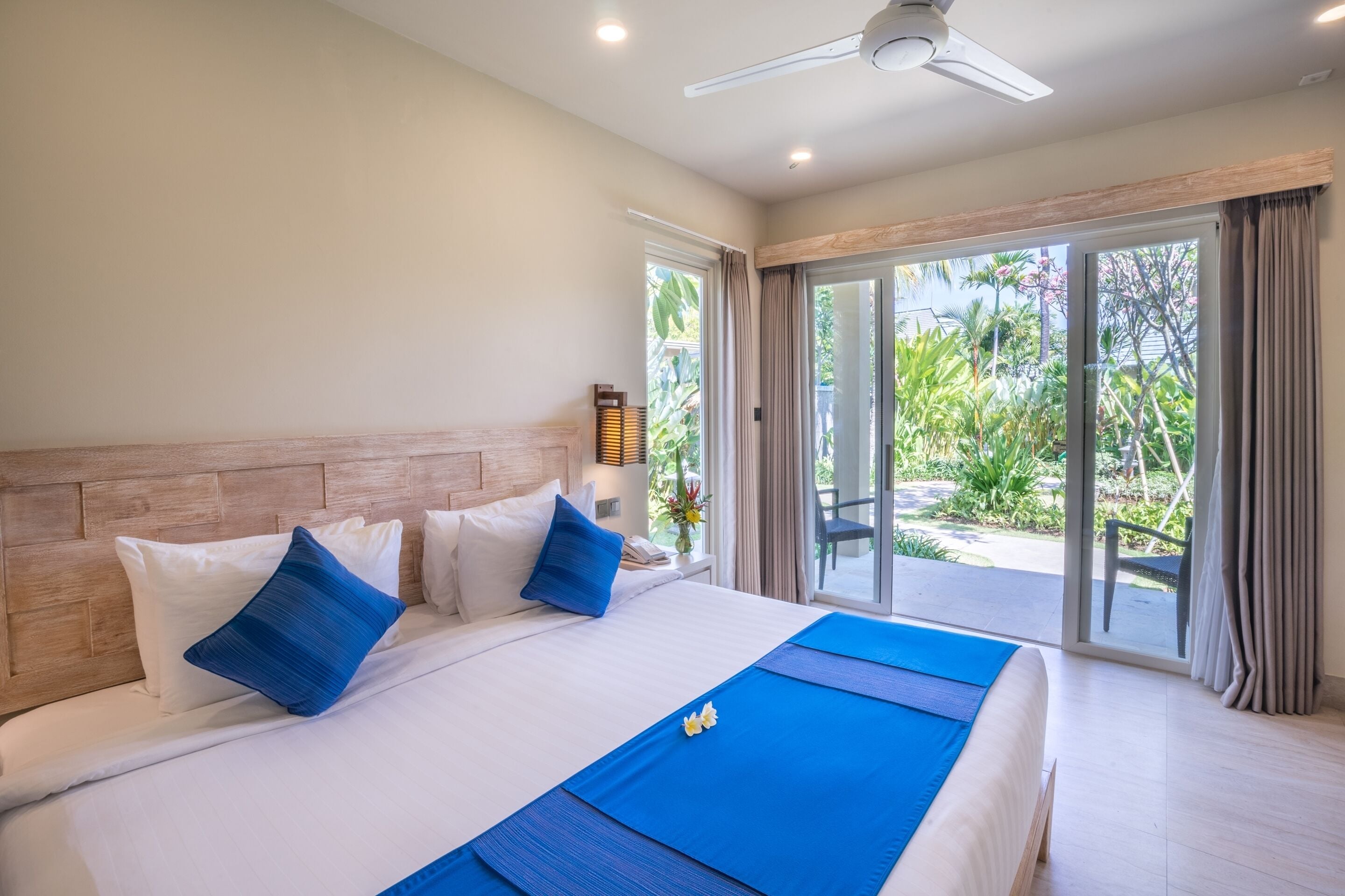 Quarto de hotel com uma cama grande, cabeceira de madeira e vista para um jardim tropical através da porta de correr de vidro.