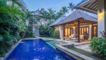 A swimming pool with a thatched-roof gazebo, surrounded by lush greenery and palm trees.