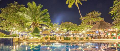 A nighttime pool area with umbrellas and lounge chairs, surrounded by palm trees and a thatched-roof structure.