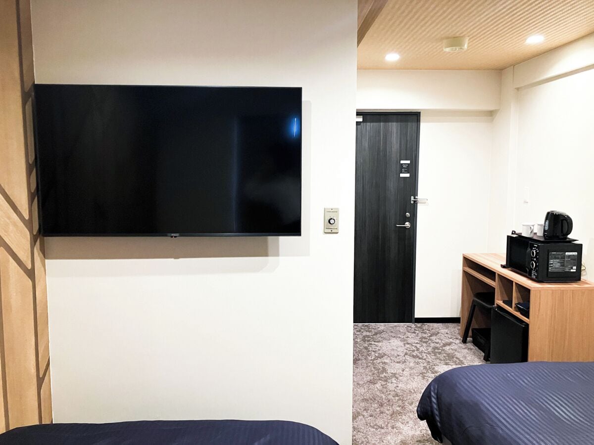 Une chambre d’hôtel dotée d’un grand téléviseur à écran plat fixé au mur, d’un bureau en bois avec un micro-ondes et de deux lits avec une literie bleu foncé.