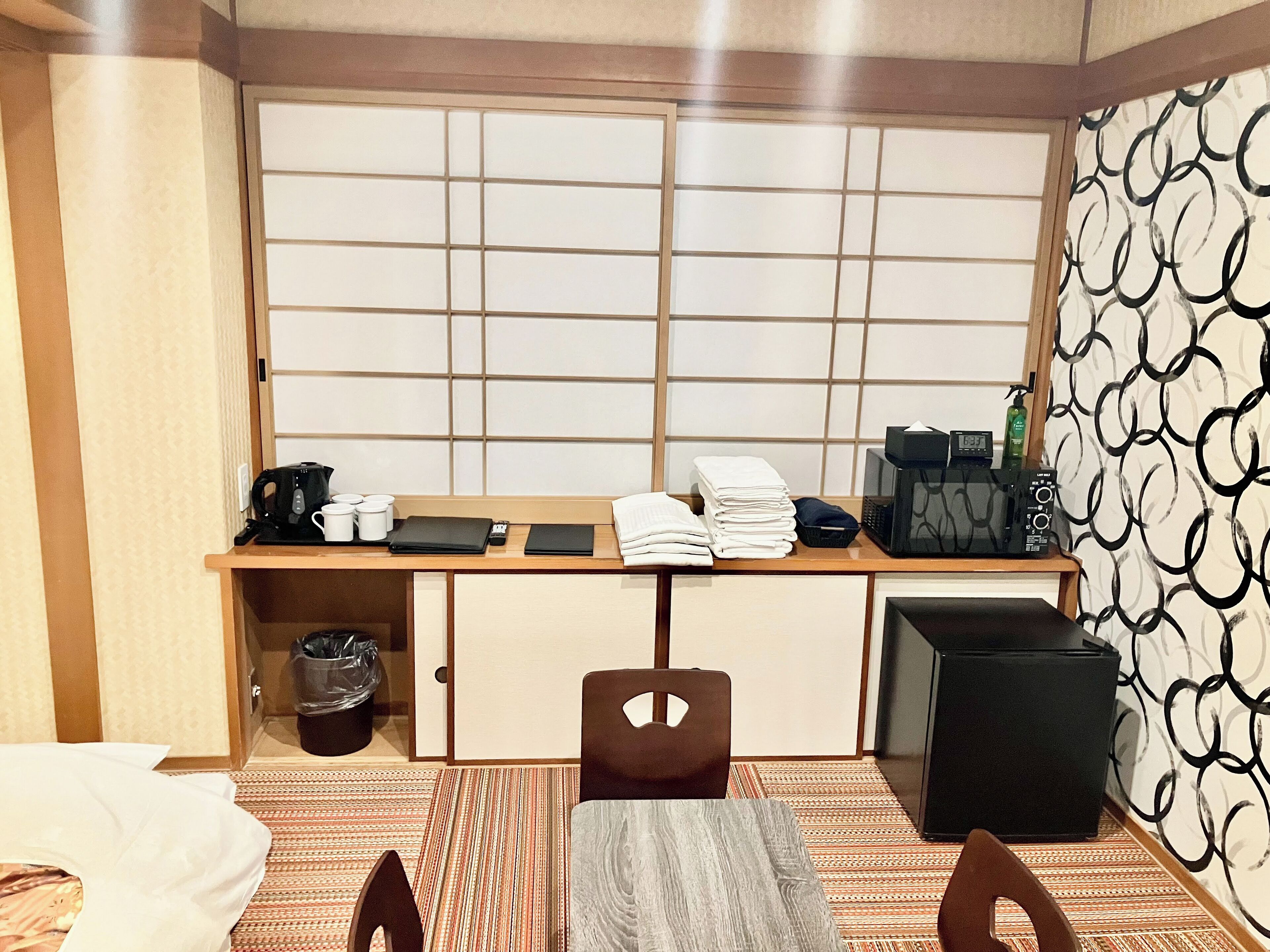 A compact hotel room with a traditional Japanese-style sliding door, a small kitchenette with a coffee maker, a microwave, and a black speaker. There is a wooden desk with a laptop, a black wallet, and a folded towel. A chair is placed at the desk, and a trash bin is visible.