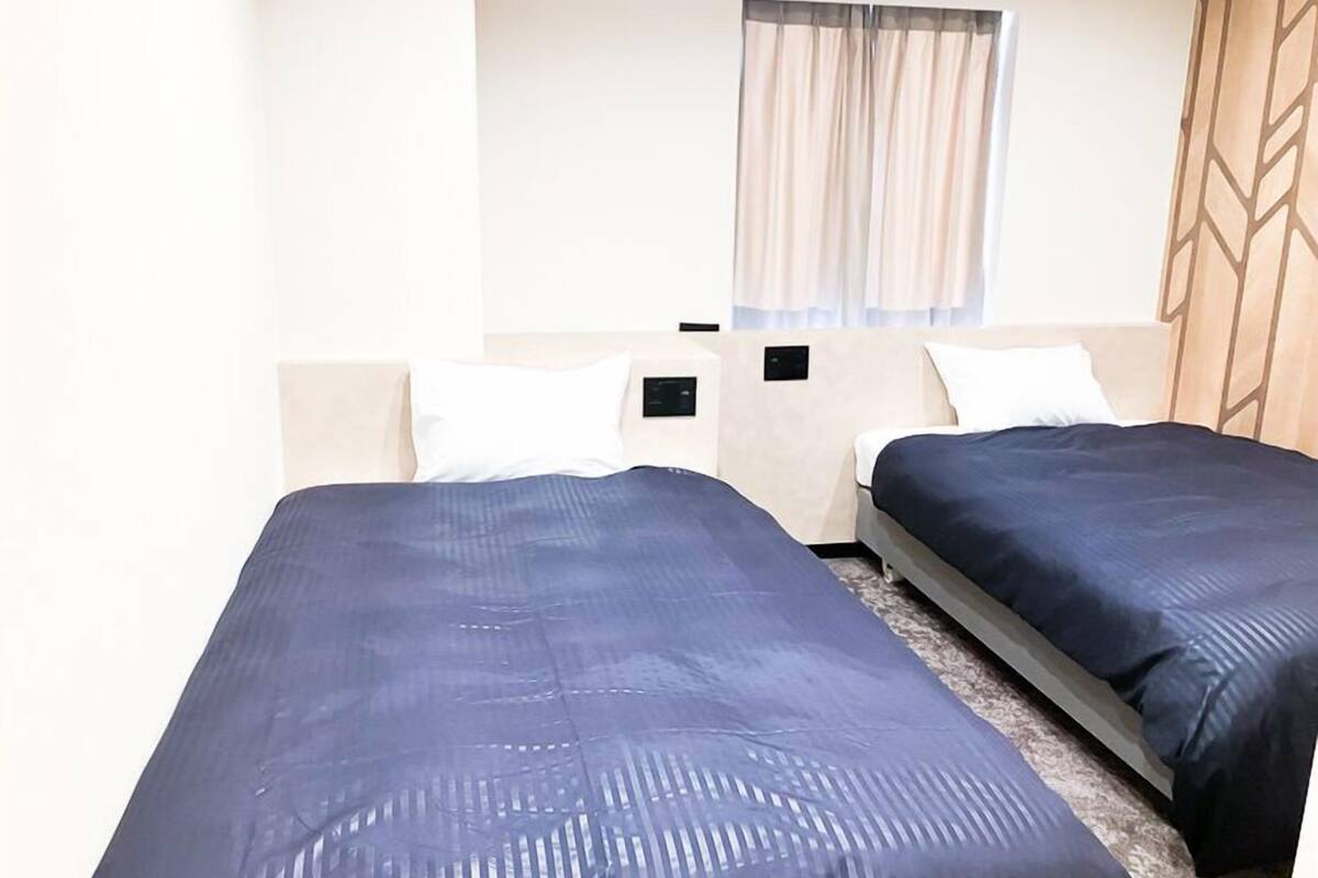 A hotel room with two beds, a window with curtains, a headboard with a geometric pattern, and a small bedside table with a lamp.