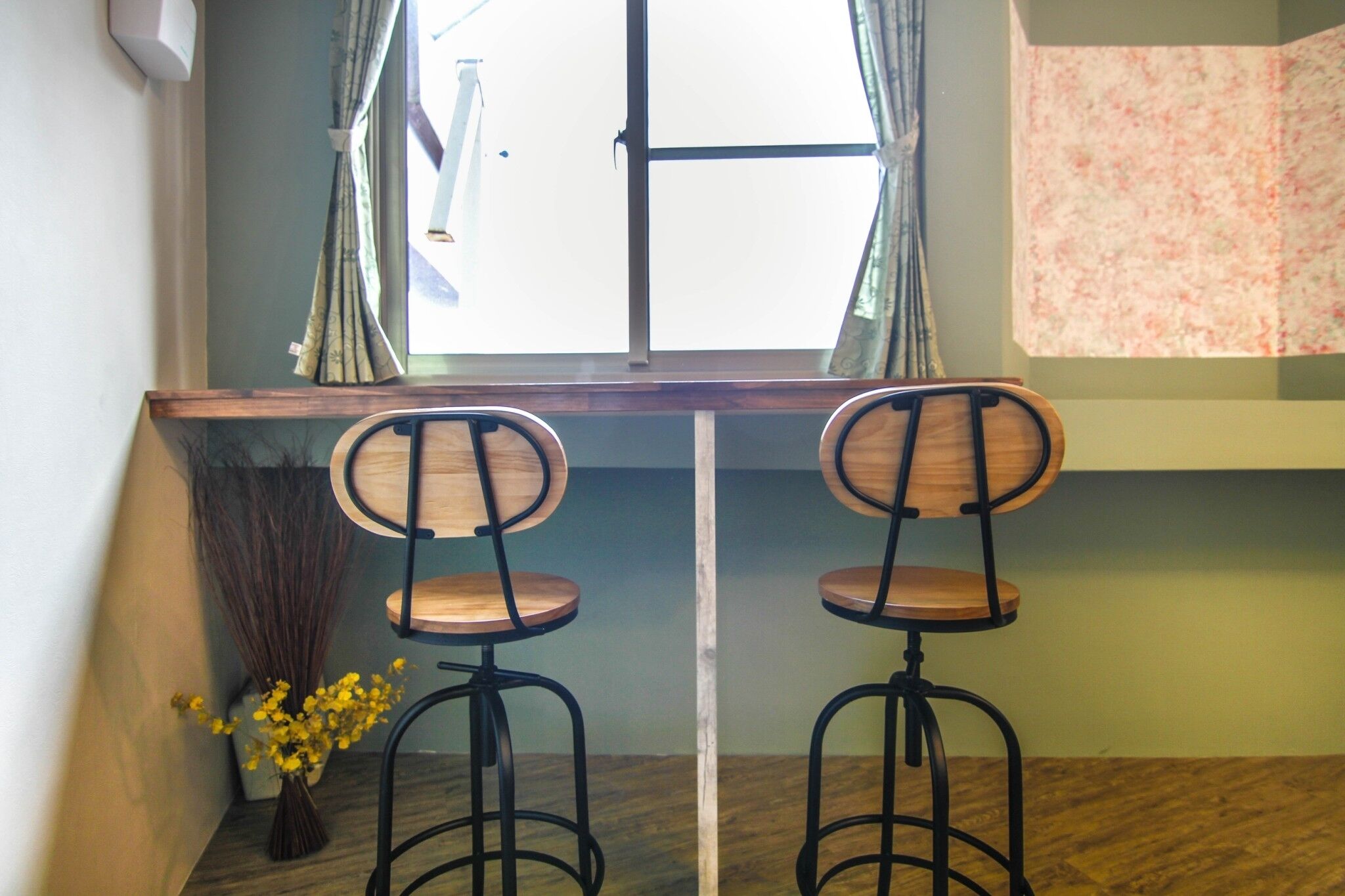 Een kamer met twee houten stoelen, een raam met gordijnen, een houten tafel en een vaas met gele bloemen.