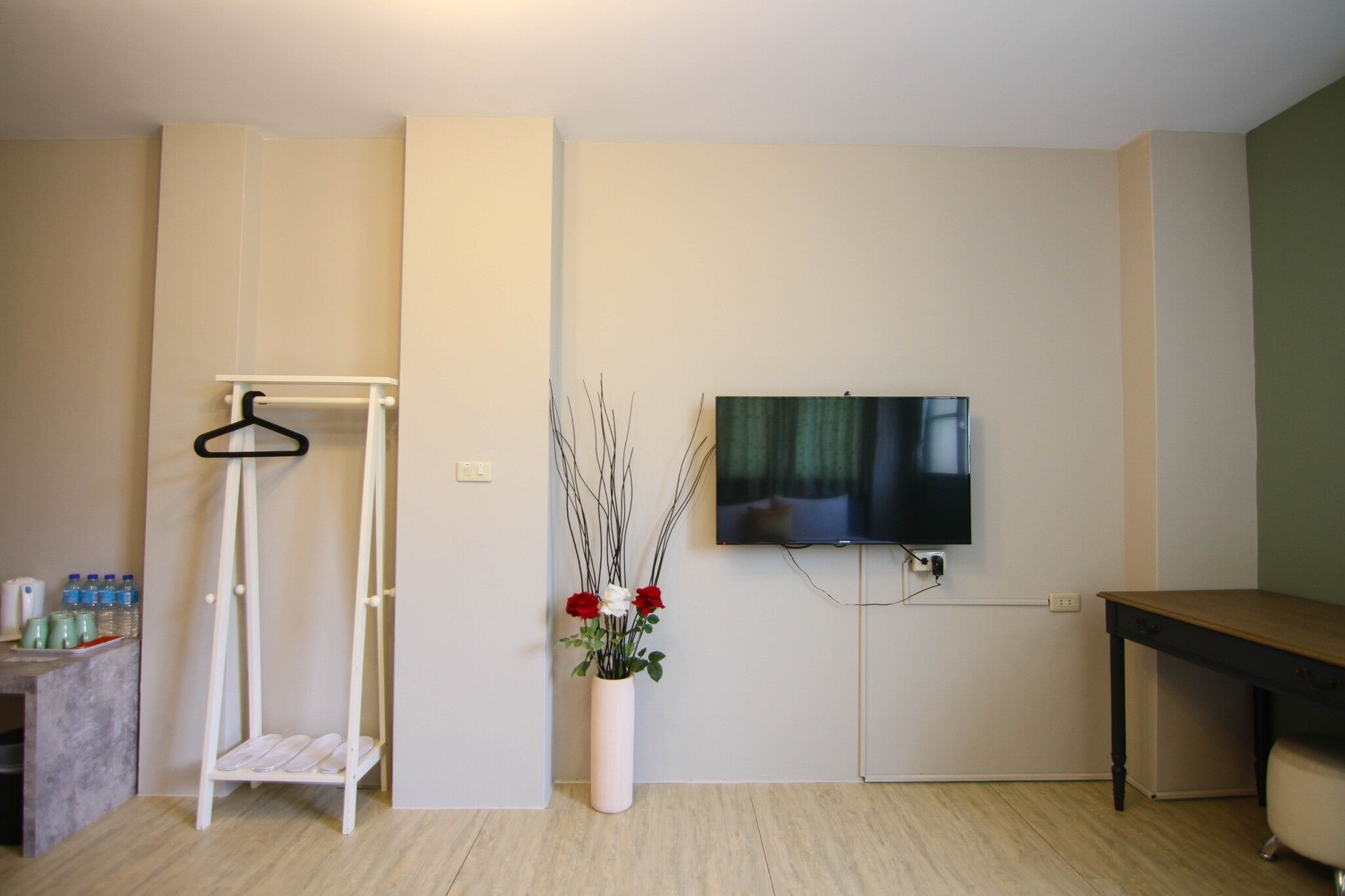 A room with a TV mounted on the wall, a white hanger stand, a vase with flowers, and a wooden table.