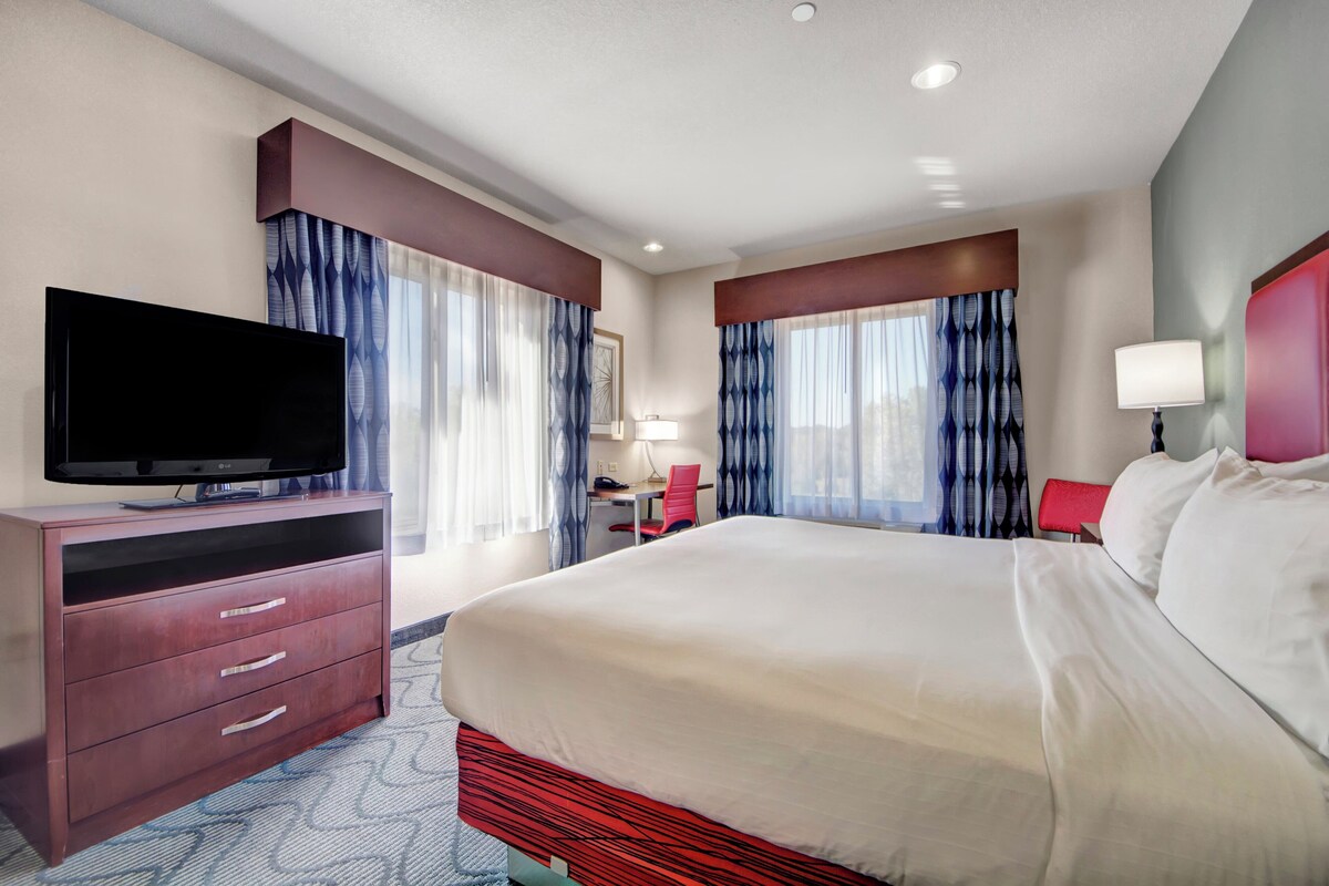 Habitación de hotel con una cama grande, un televisor sobre un aparador de madera, un escritorio con silla y ventanas amplias con cortinas.