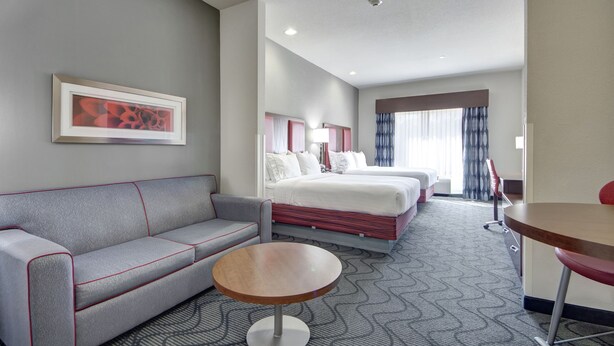 A hotel room with a grey sofa, a round table, a bed with white linens, and a framed picture on the wall.