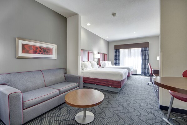 Una habitación de hotel con un sofá gris, una mesa redonda, una cama con ropa blanca y un cuadro en la pared.