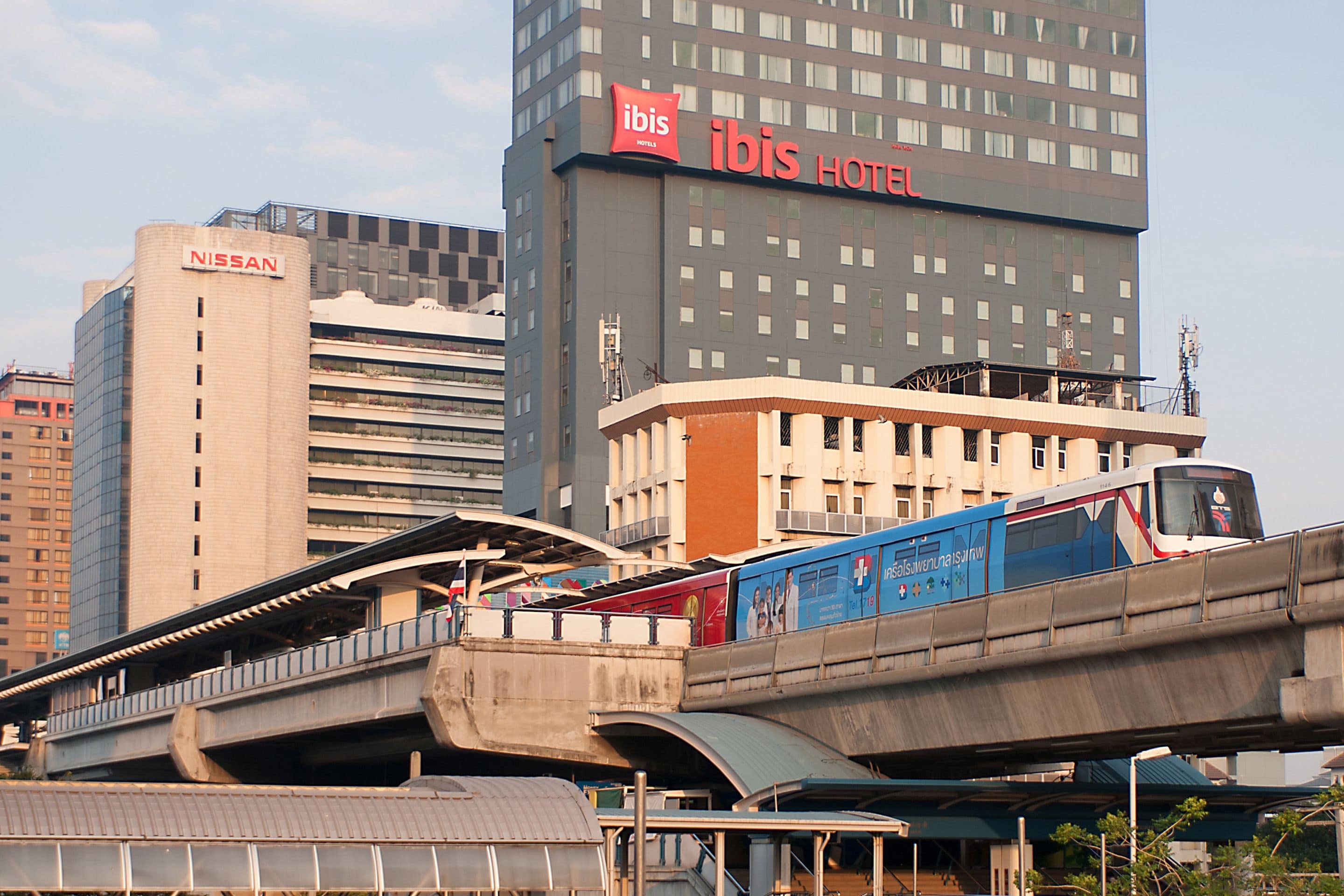 A modern hotel with a prominent "Ibis Hotel" sign, a Nissan building, and a train passing over an elevated walkway.