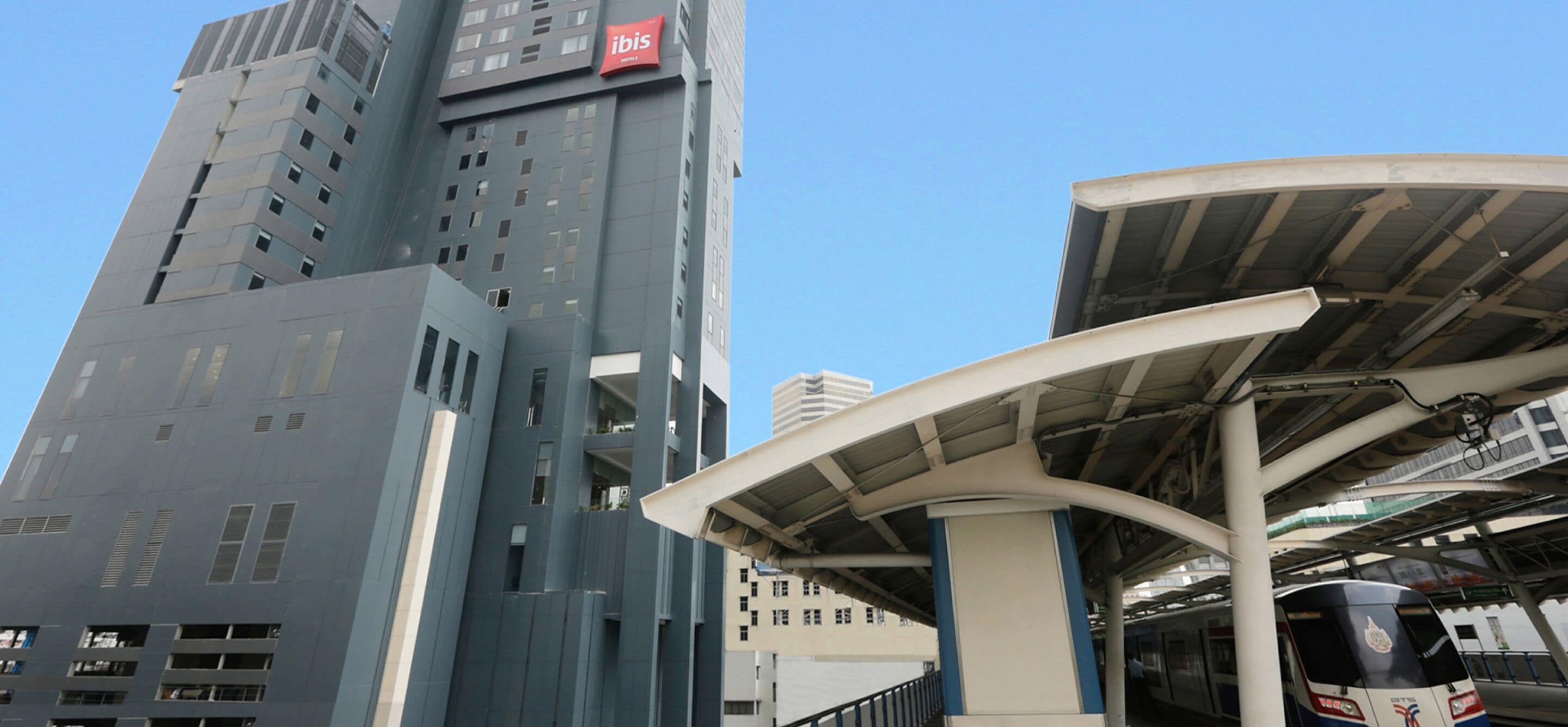 A modern building with a prominent 'ibis' sign, a covered waiting area, and a train platform.