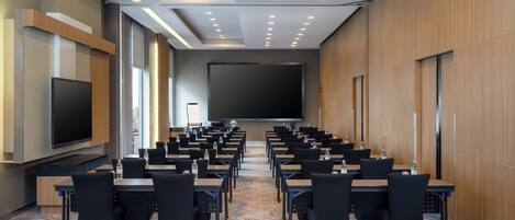 Un salón de conferencias con filas de mesas y sillas, una pantalla grande y un proyector.