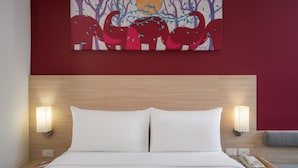 A hotel room with a wooden headboard, white bedding, two bedside lamps, a telephone, and a painting above the bed. - ibis Bangkok Siam (Bangkok)