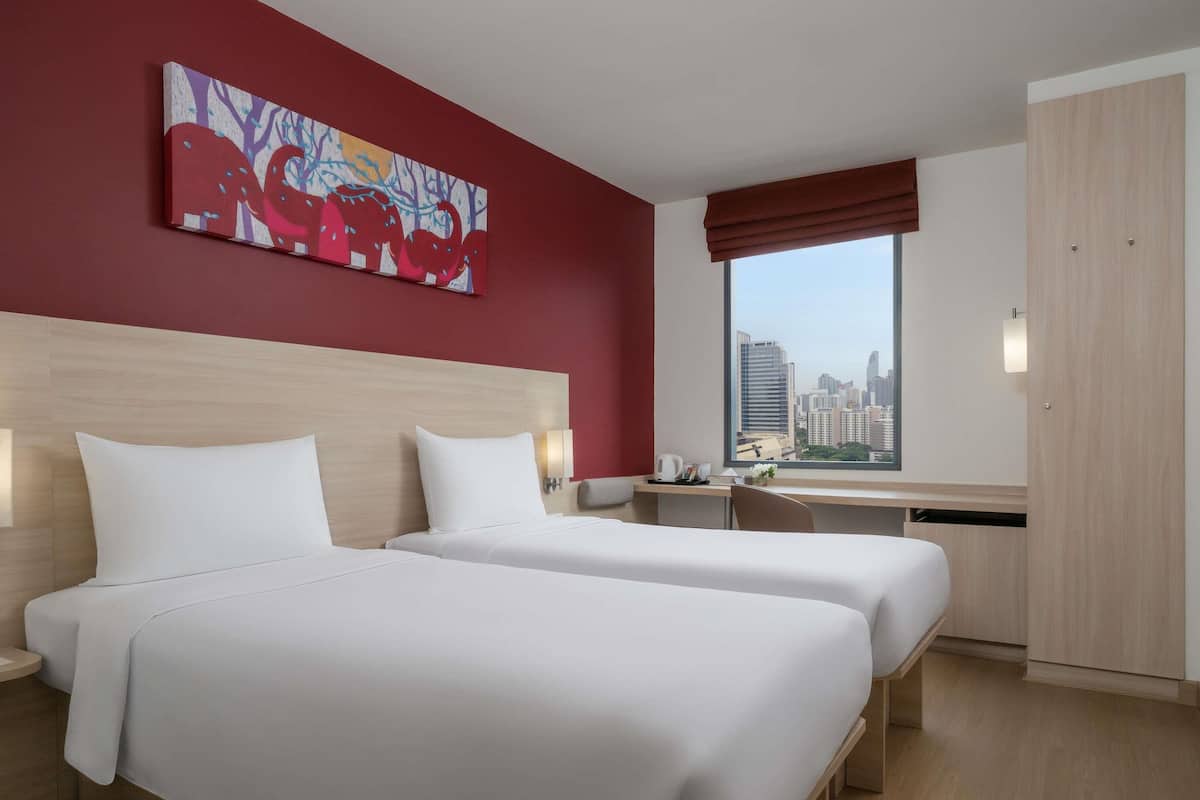Habitación de hotel con dos camas, un escritorio, una ventana con vistas a la ciudad y un cuadro abstracto y colorido en la pared.