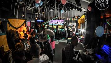 Patrons gather in a lively bar area with hanging decorations and a brick wall.