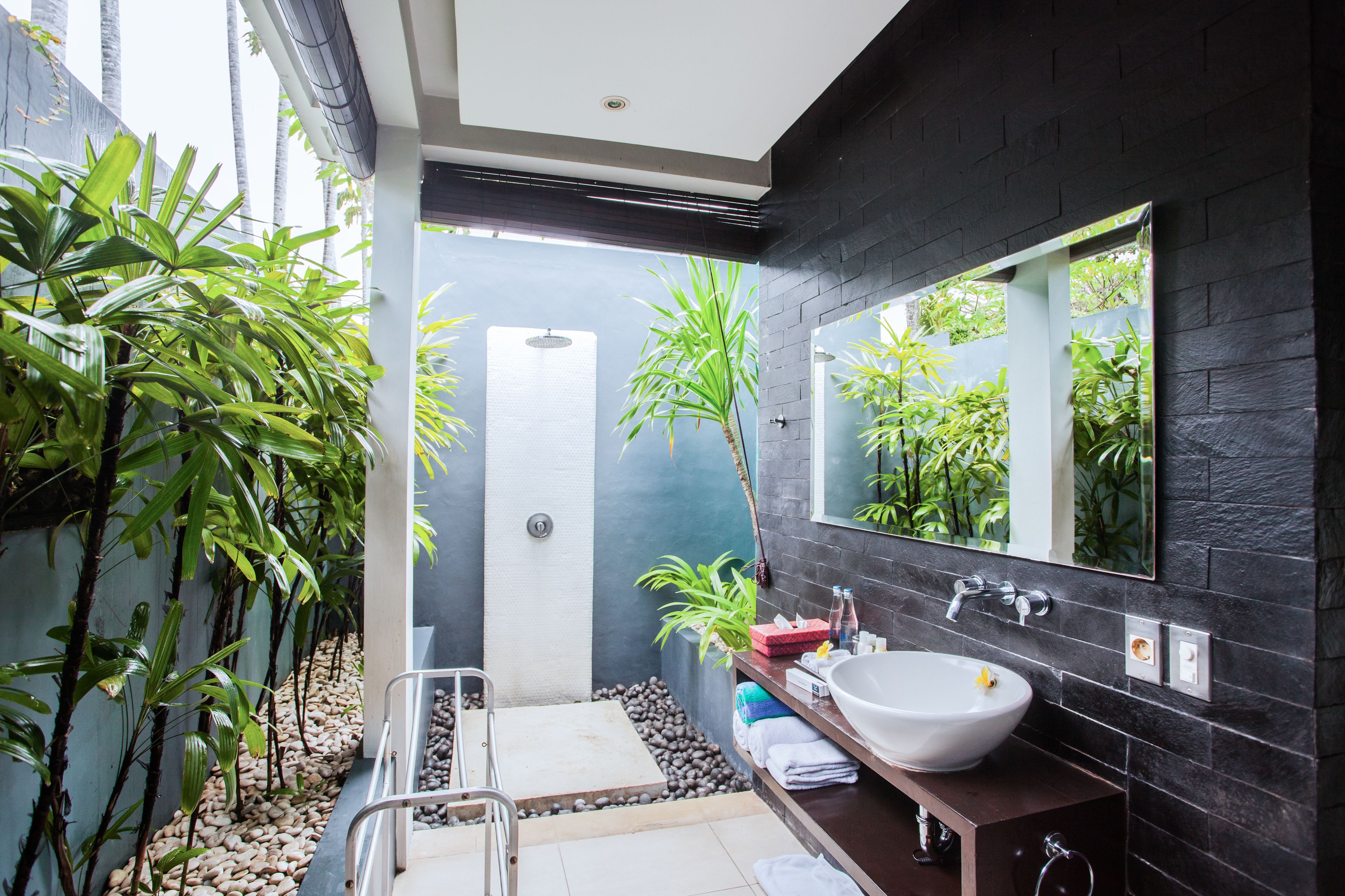 A modern bathroom with a large mirror, white sink, and black tiled walls. There are plants and a small shower area visible.