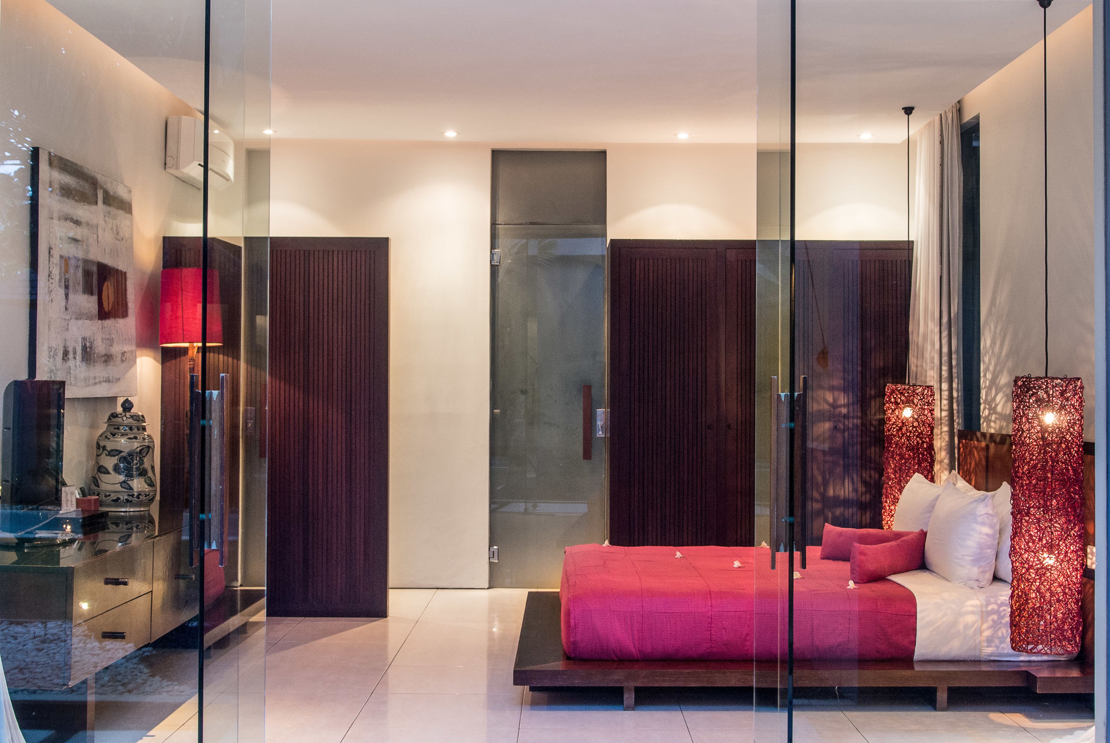 A modern bedroom with a glass partition, a red bed, and a nightstand with a lamp.