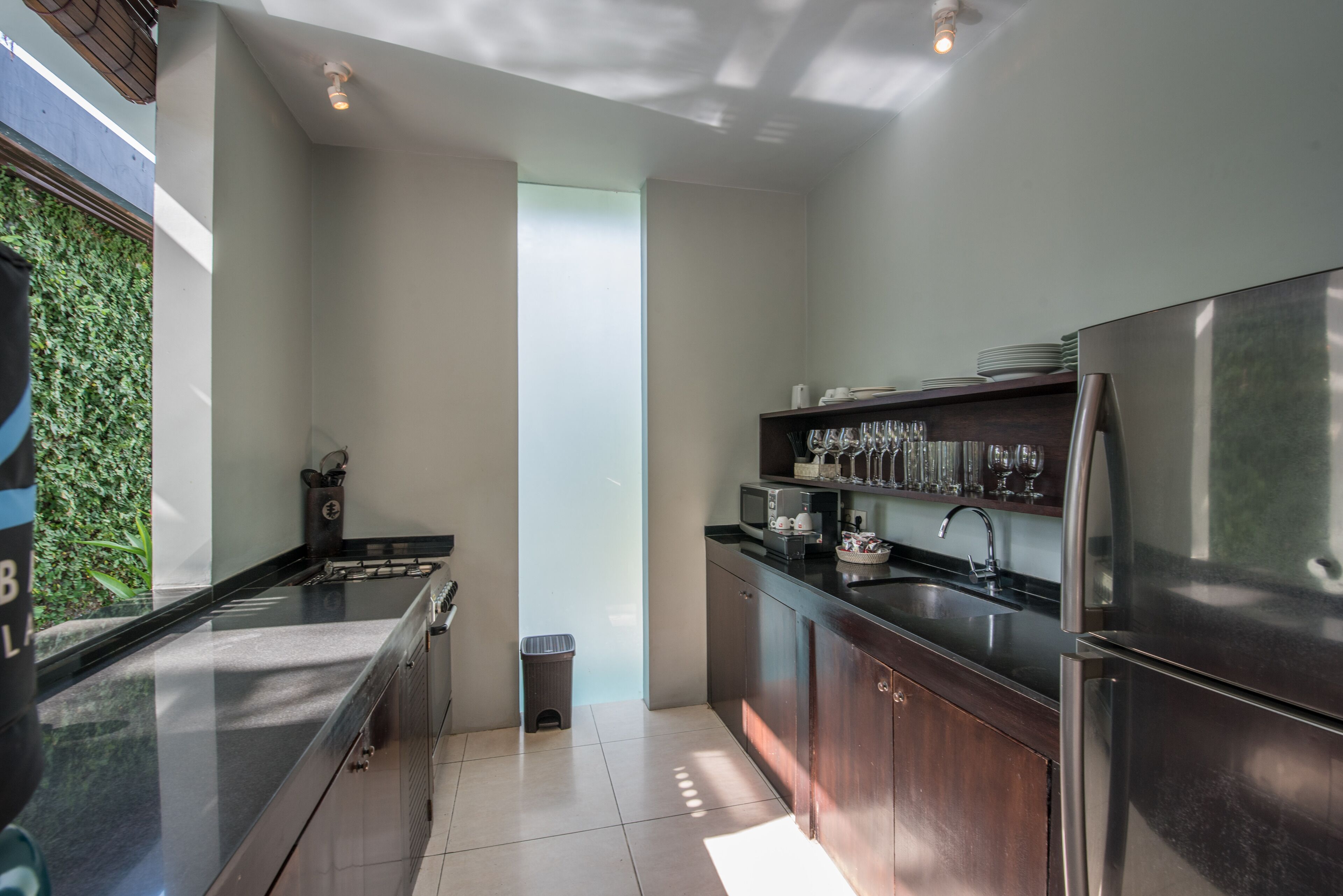 A modern kitchen with a stainless steel refrigerator, a microwave, and a sink. There is a trash can and a window with a view of greenery.
