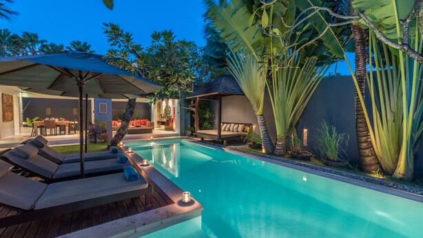 A swimming pool with lounge chairs and a covered seating area, surrounded by tropical plants. - Kembali Villas (Seminyak)