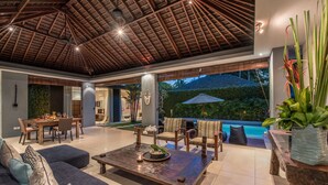 A spacious indoor area with a wooden ceiling, a pool visible through the glass wall, and a dining area with a table and chairs. - Kembali Villas (Seminyak)