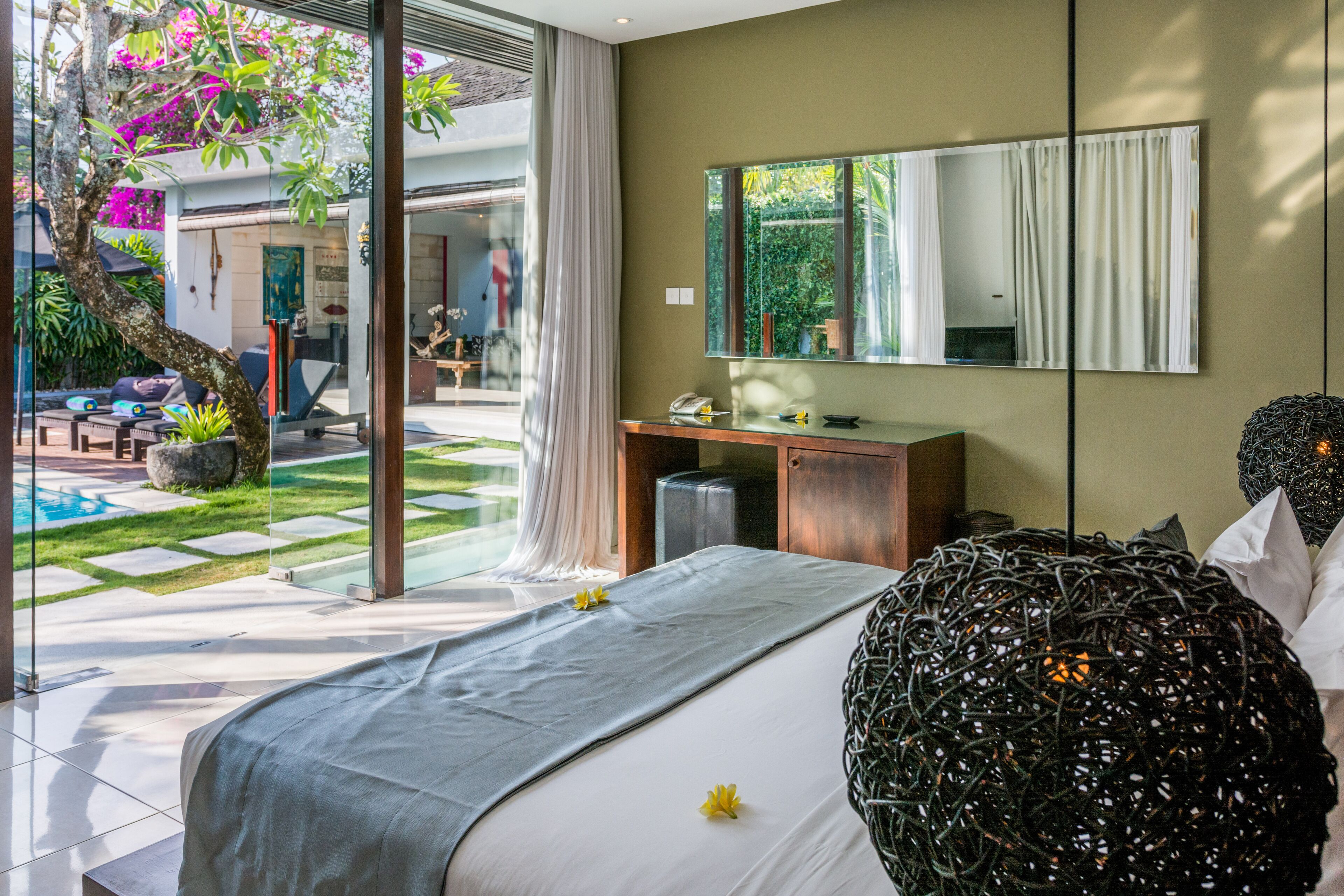 A modern bedroom with a large bed, a desk, and a mirror. There is a glass door leading to a garden with a pool and outdoor furniture.