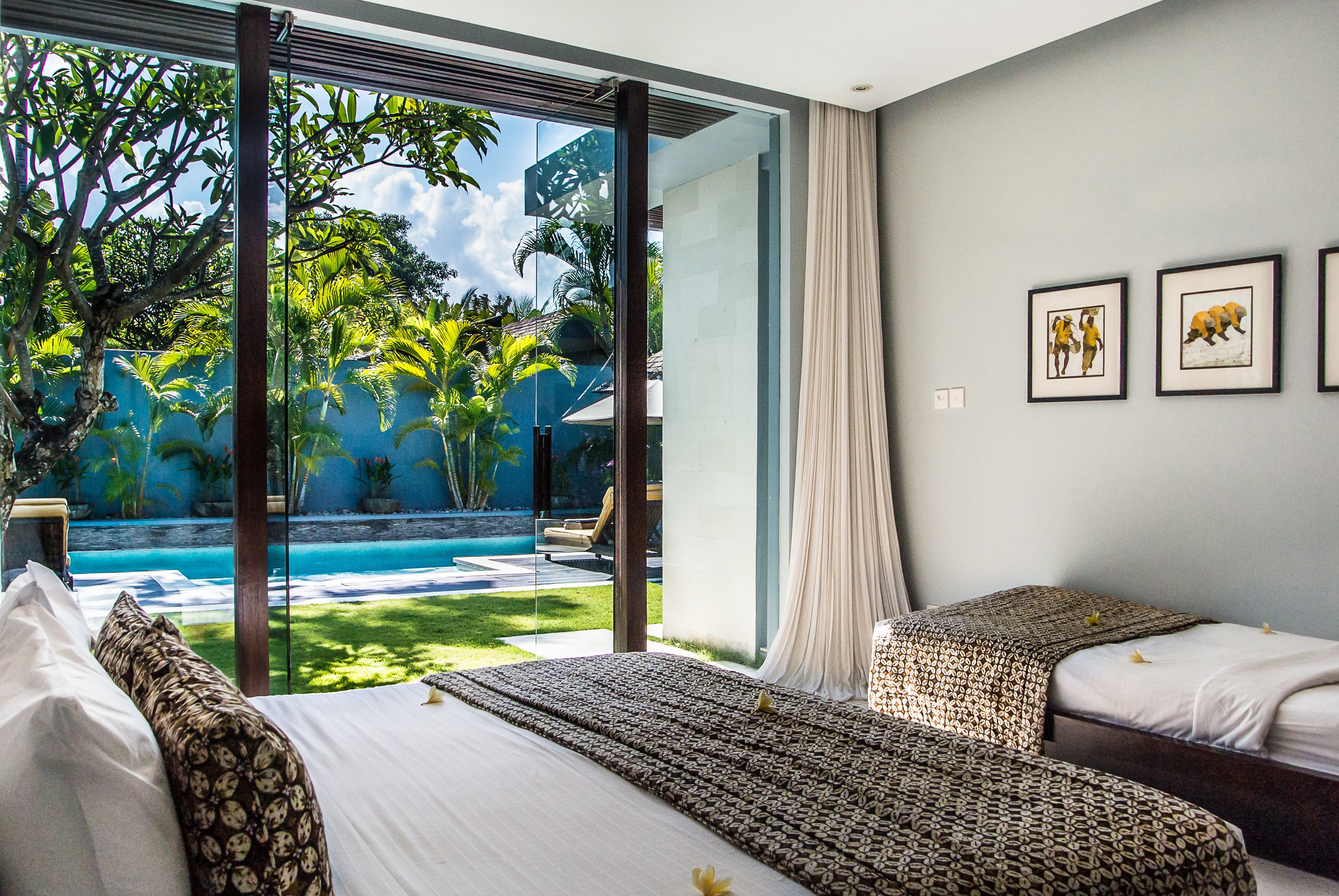 A hotel room with two beds, a large window with a view of a swimming pool and tropical garden, and framed artwork on the wall.