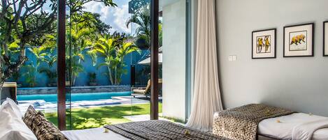 A hotel room with two beds, a large window with a view of a swimming pool and tropical garden, and framed artwork on the wall.