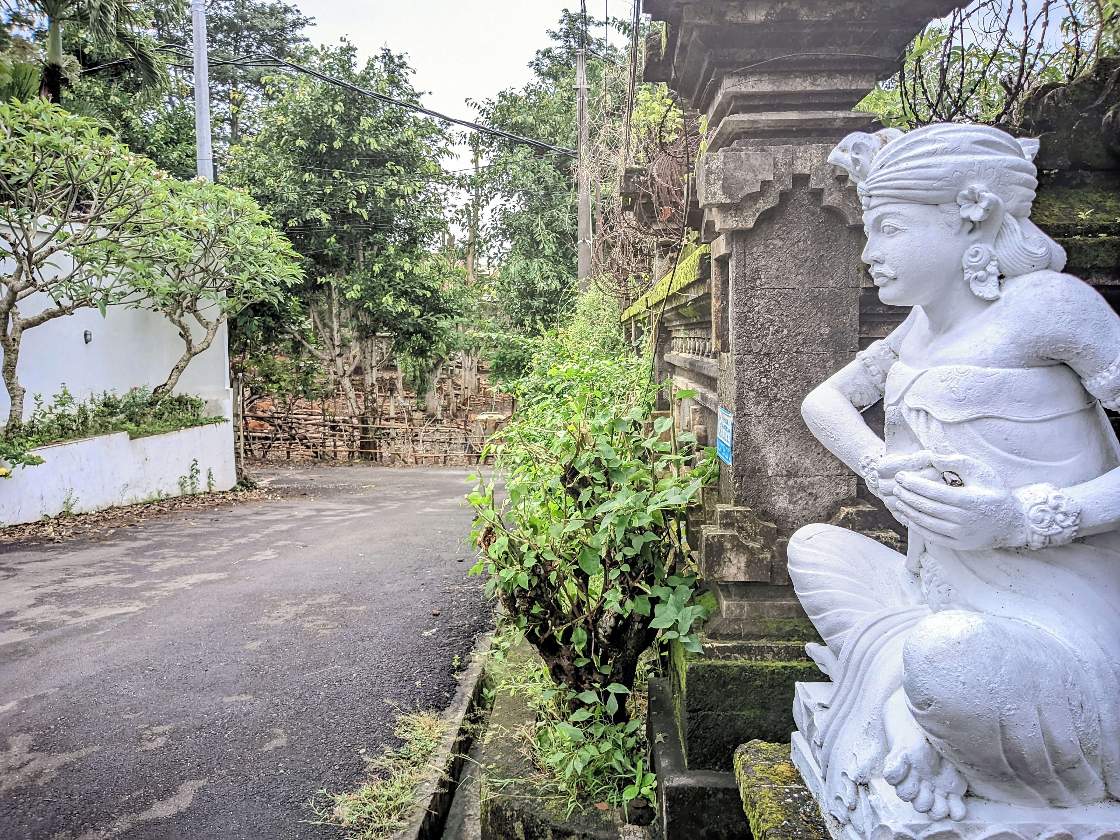 Una estatua de una figura sentada con un rostro y manos detallados, frente a un fondo de follaje y una estructura de piedra.