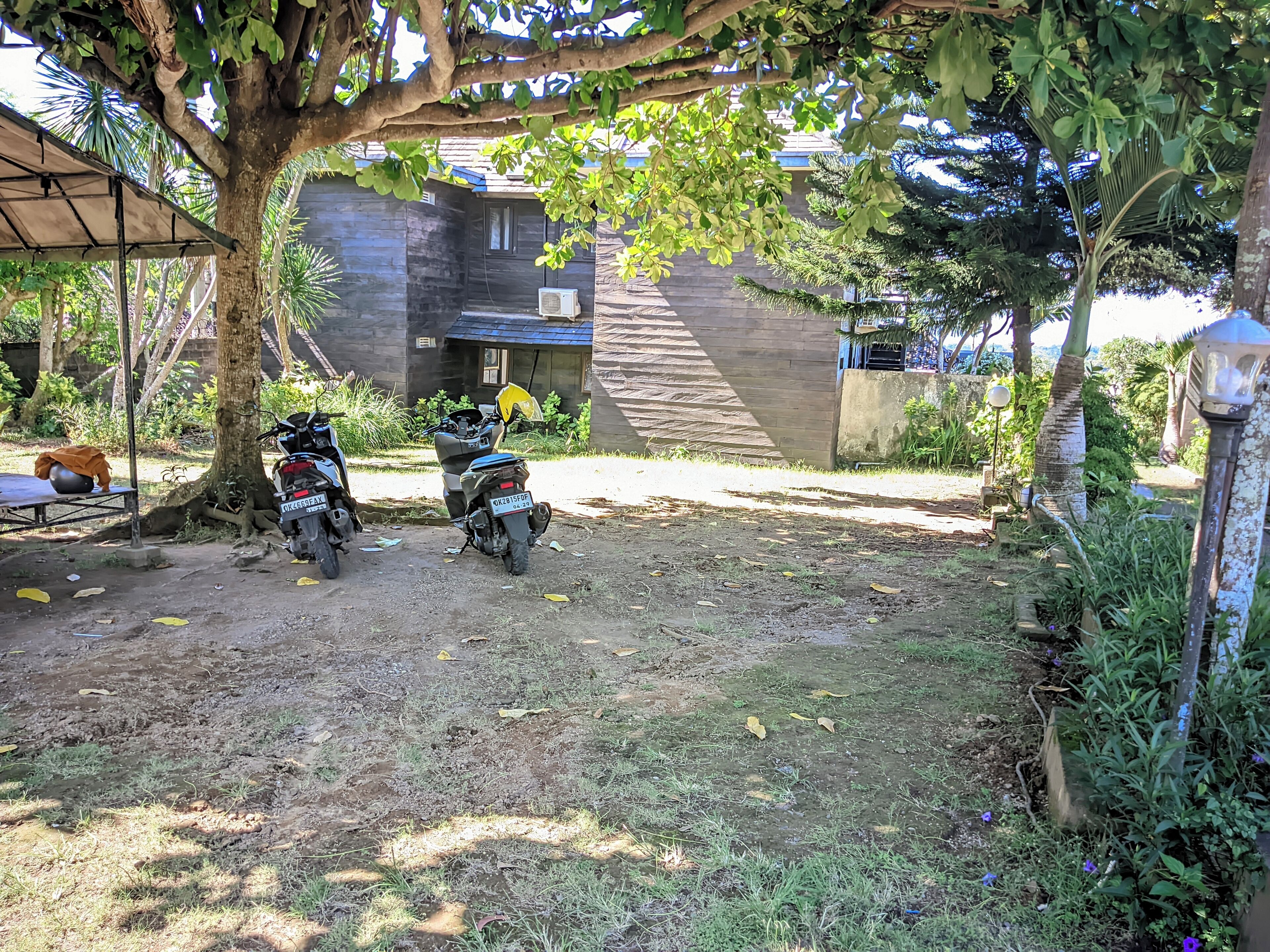 Un espacio al aire libre cubierto con dos motocicletas estacionadas bajo un árbol, rodeado de vegetación y un edificio con techo azul.