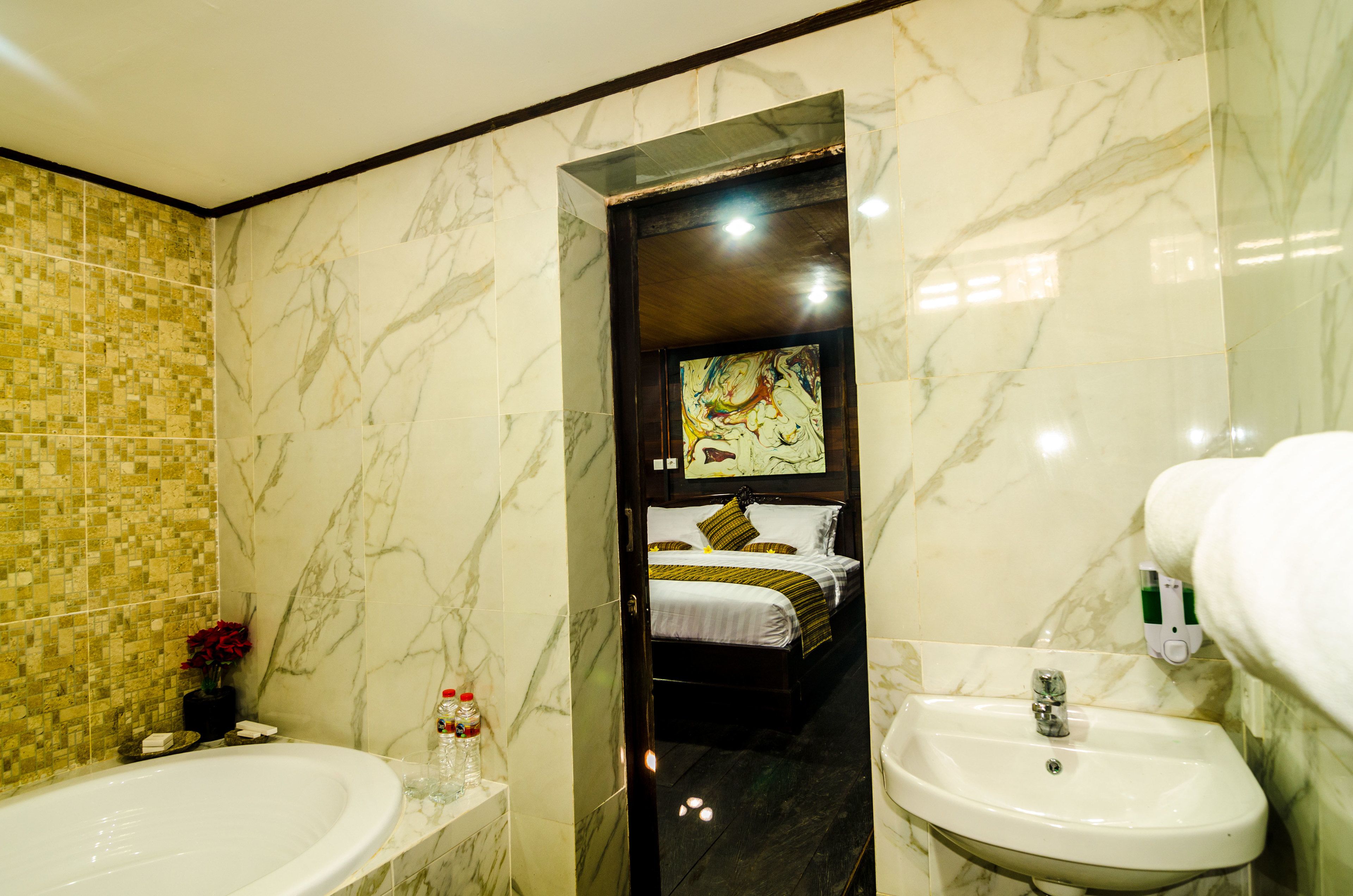 A bathroom with a marble-tiled wall, a bathtub, a sink, and a bed visible through a doorway.