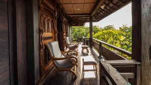 A wooden porch with ornate carvings, a row of cushioned chairs, and a view of lush greenery. - The Kawan Jimbaran (Ungasan)